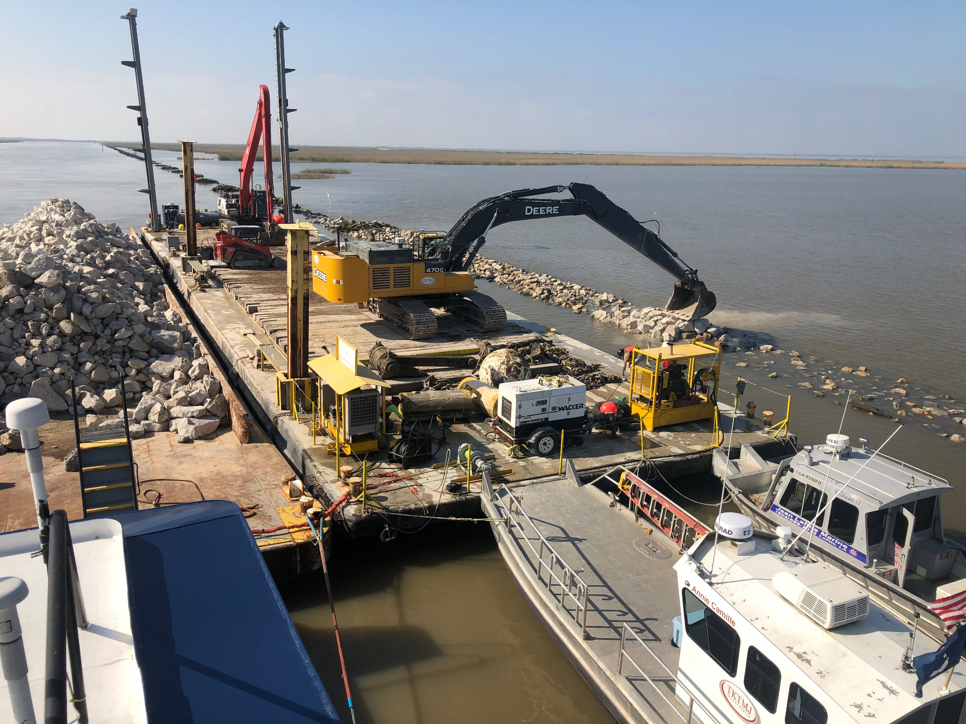 Excavator on a deck barge placing riprap along a rock breakwater, with TKTMJ crew boats alongside