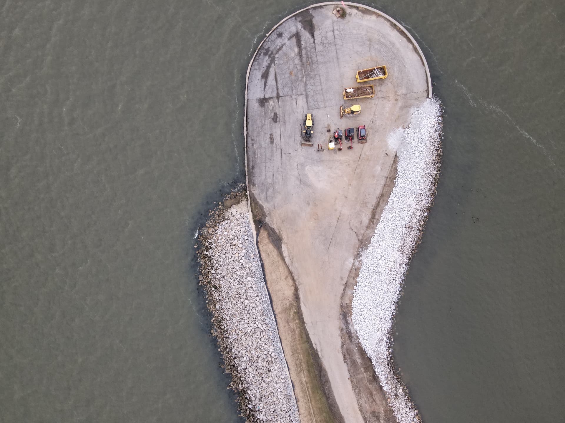 Aerial view of a rock breakwater curving around a point of land, with equipment staged on shore